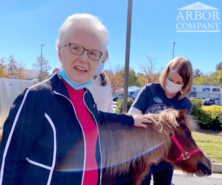 A resident joyfully pets a small horse outdoors on a sunny autumn day with a staff member nearby, showcasing the unique, enriching experiences that define wellness-focused senior living East Cobb and senior care Marietta GA.