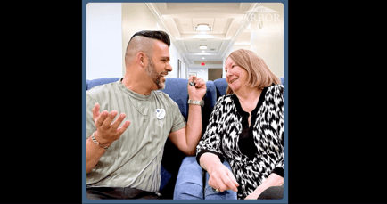 A staff member and a senior woman share a lively, joyful conversation while seated in a hallway, capturing the warm community bonds of the Evergreen Neighborhood at Arbor Terrace Mountainside NJ.