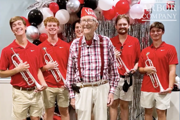 A smiling older man in a red paper Varsity hat, red-and-white plaid shirt, and maroon suspenders stands at the center of a celebration. He is surrounded by five young men in red Georgia polo shirts, each holding a silver trumpet, in front of a backdrop of red, black, white, and silver balloons and silver tinsel.