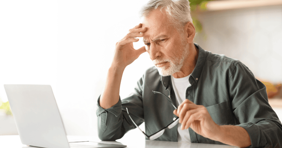 An older man with gray hair and a beard sits at a table, holding his glasses in one hand and pressing the other to his forehead in a worried or frustrated expression while looking at a laptop, with a printed document on the table in front of him.