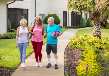 Three older adults in athletic wear walk together along a landscaped outdoor pathway, laughing and smiling. Two women and one man carry rolled yoga mats, suggesting they are heading to or returning from a fitness class. The background features a well-maintained building with manicured hedges, a palm tree, and lush green lawn