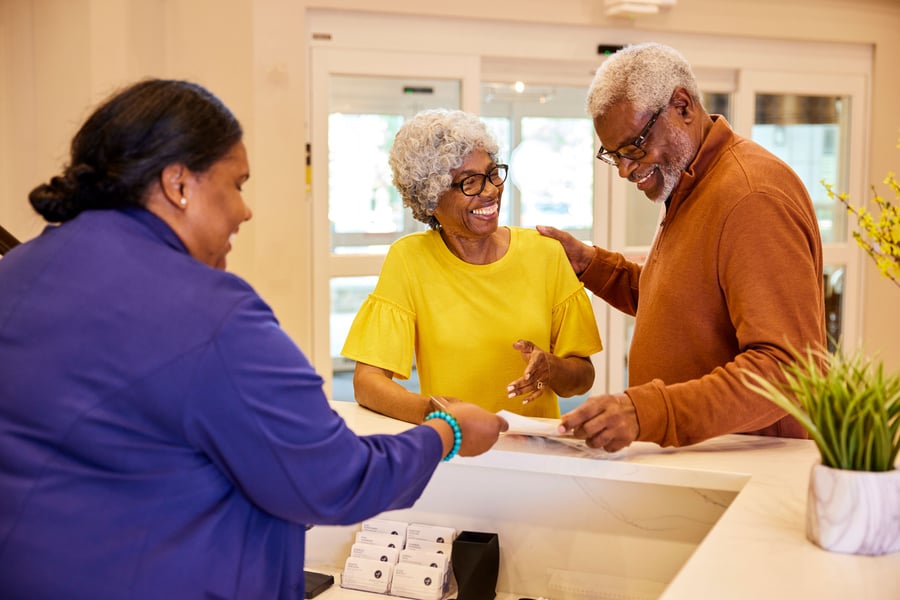 An older couple smiling at a front desk while a staff member in a purple jacket hands them paperwork, with business cards and a small plant on the counter.