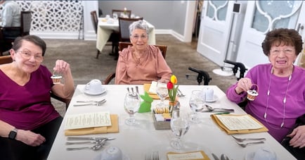 Three senior women smile and hold small treats at a beautifully set brunch table adorned with tulips and glassware, enjoying a delightful social dining experience at Arbor Terrace Mountainside NJ.