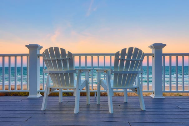 Two white Adirondack chairs face a calm ocean at sunset from a wooden deck, evoking the peaceful, coastal lifestyle enjoyed by seniors living in Fort Myers, FL.