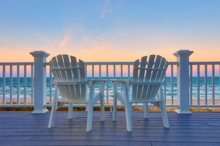 Two white Adirondack chairs face a calm ocean at sunset from a wooden deck, evoking the peaceful, coastal lifestyle enjoyed by seniors living in Fort Myers, FL.