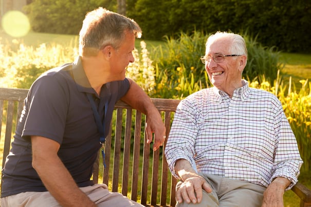 A staff member and an older male resident share a warm conversation on a wooden bench surrounded by golden sunlight and lush garden blooms, embodying the nature-access senior living Woodbridge VA community.