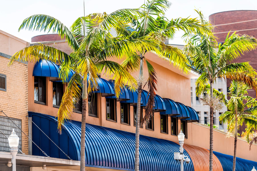 A sunny street view in Fort Myers, FL, featuring a warm-toned building with blue awnings and tall swaying palm trees, reflecting the vibrant community surrounding senior living in the area.
