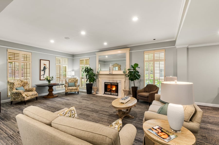 A bright and inviting common area at a senior living community in Athens, GA, featuring a lit fireplace, patterned armchairs, neutral sofas, lush greenery, and large plantation-shuttered windows filling the space with natural light.
