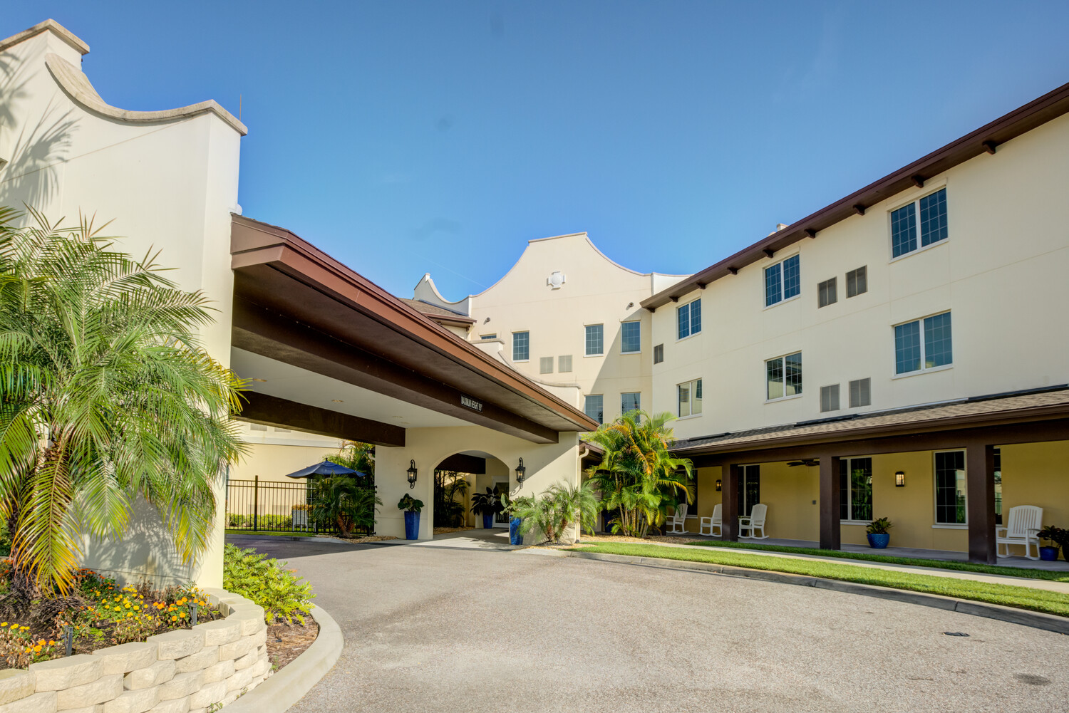 Inviting porte-cochère entrance with palm trees and rocking chairs, showcasing the resort-style senior living in Citrus Park, FL.