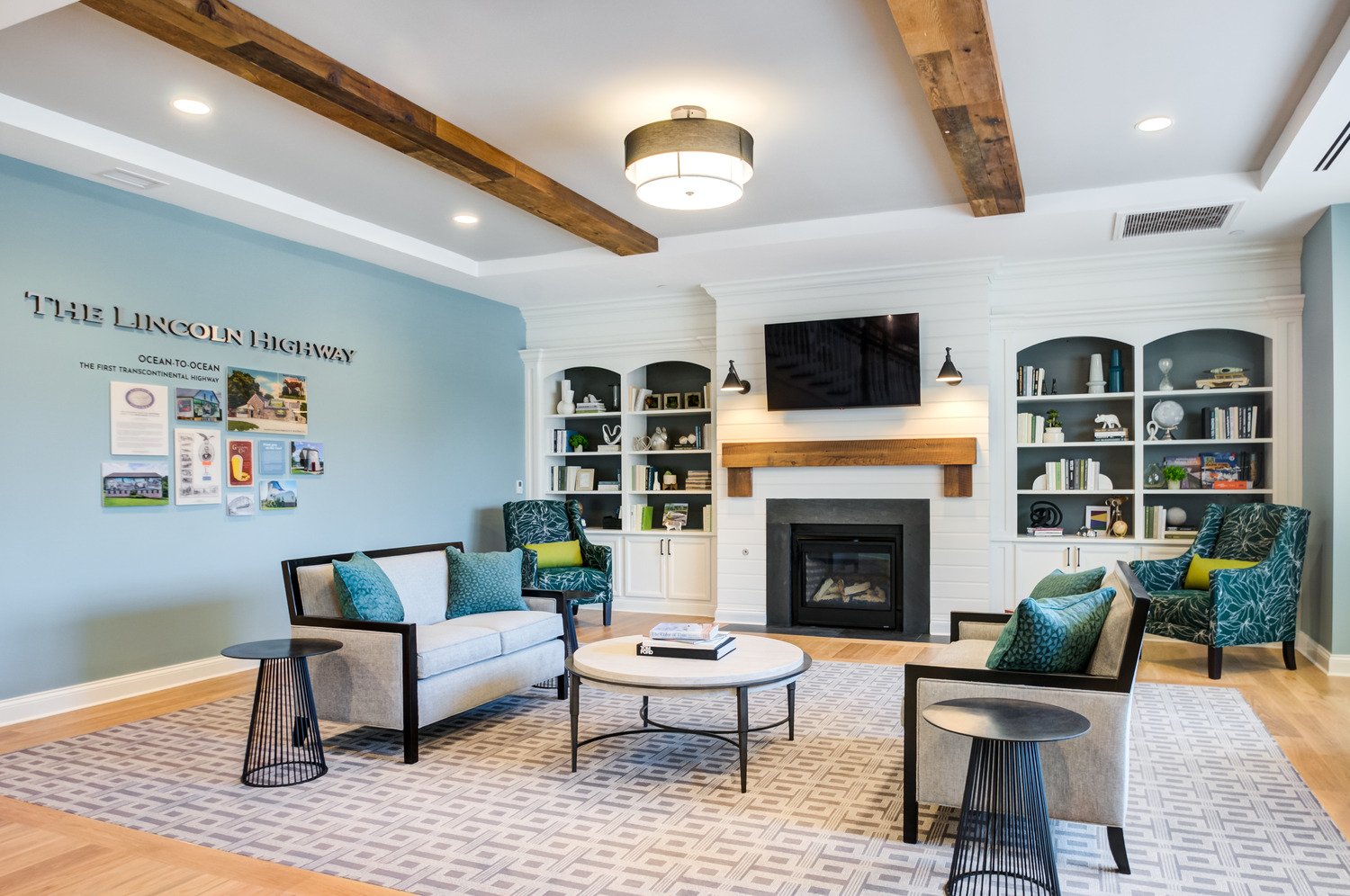 Arbor Terrace Exton common spack with wooden floors and a grey rug. grey couches with blue pillows on each side of a coffee table. two blue chairs with yellow pillows. a blue wall with artwork and a white wall with cubby shelves and a fireplace with tv. 