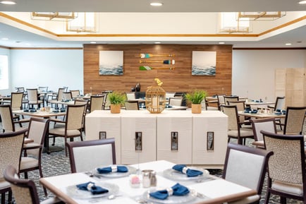 A well-appointed dining room at Barrington Terrace of Fort Myers, FL, featuring neatly set tables, warm wood accent walls, coastal-themed artwork, and soft neutral tones throughout the inviting space.