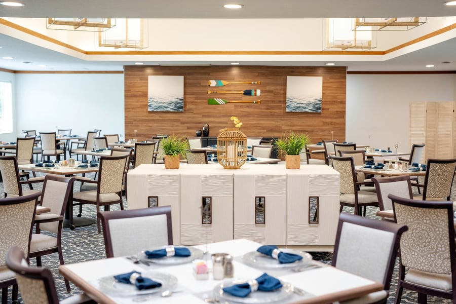 A well-appointed dining room at Barrington Terrace of Fort Myers, FL, featuring neatly set tables, warm wood accent walls, coastal-themed artwork, and soft neutral tones throughout the inviting space.