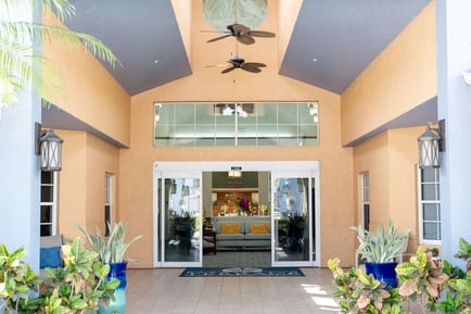 The bright, welcoming exterior entrance of Barrington Terrace of Fort Myers features a vaulted ceiling, ceiling fan, lantern sconces, and lush green plants flanking open glass doors.
