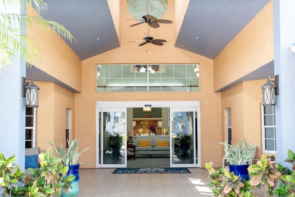 The bright, welcoming exterior entrance of Barrington Terrace of Fort Myers features a vaulted ceiling, ceiling fan, lantern sconces, and lush green plants flanking open glass doors.
