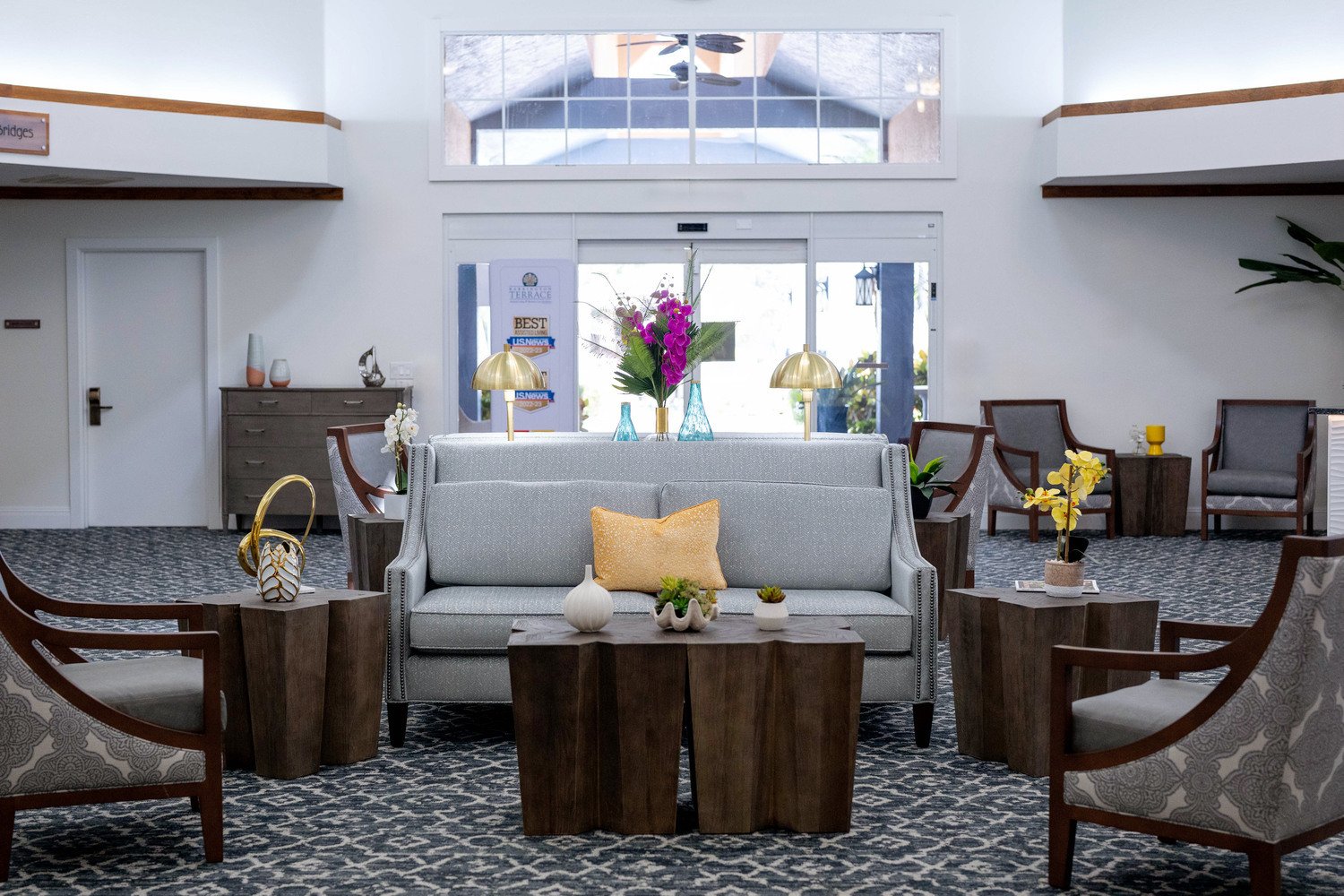 Fort Myers lobby area with a two grey and dark doowned chairs and a couch with a yellow pillow surrounding a coffee table. Big entryway with windows.
