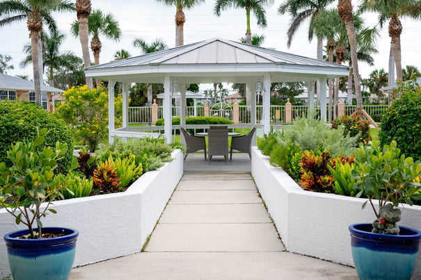 The welcoming entrance of Barrington Terrace of Fort Myers features warm peach-toned walls, lantern sconces, lush potted plants, and open glass doors revealing a cozy interior seating area.