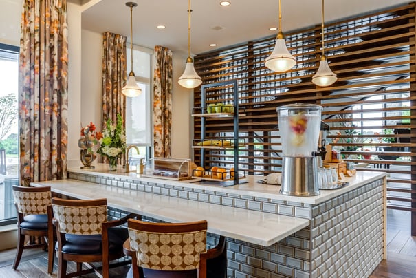 A bright refreshment bar features fresh fruit, infused water, and elegant tiled accents beneath statement pendant lighting, reflecting the senior living with wellness programs Fulton MD and senior living near Jessup MD offer.