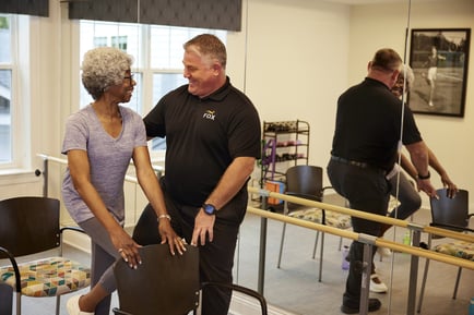 A resident and a fitness staff member share a cheerful laugh during a wellness session in a bright exercise room with ballet barres and mirrors, reflecting the active senior community life at Prince William County assisted living.