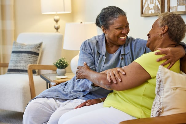 Two older women share a joyful, heartfelt laugh while embracing on a couch in a cozy, well-lit living space, reflecting the warm friendships and vibrant social activities that define senior living Woodbridge VA.
