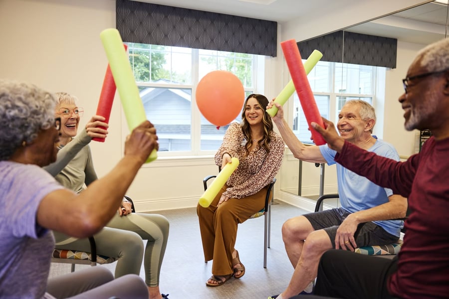 A cheerful group enjoys a lively balloon and pool noodle activity together indoors, highlighting the fun wellness programs at a senior living community in Suwanee, GA.