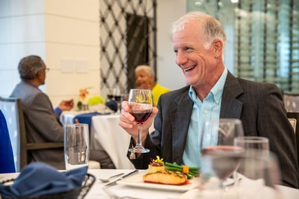 A well-dressed older male resident smiles warmly while raising a glass of red wine over a plated meal in an upscale dining room, capturing the resort-style senior living atmosphere in Woodbridge, VA.