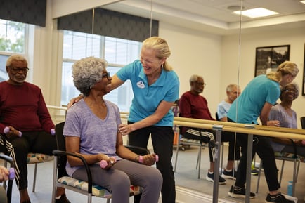 A staff member smiles warmly with a resident during a seated fitness class at our senior living East Cobb community, as others exercise with hand weights in a bright, welcoming activity room.