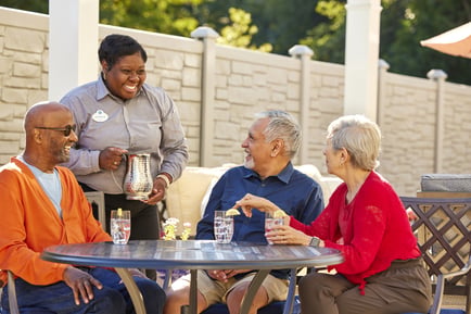 A smiling staff member serves refreshments to residents enjoying conversation at an outdoor patio table, highlighting the attentive, personalized assisted living East Cobb experience at our senior living Marietta GA community.