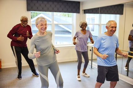 A group of seniors laughs and moves together during a lively dance fitness class in a bright studio — the active senior lifestyle that defines the Arbor Terrace Lakeway TX difference.