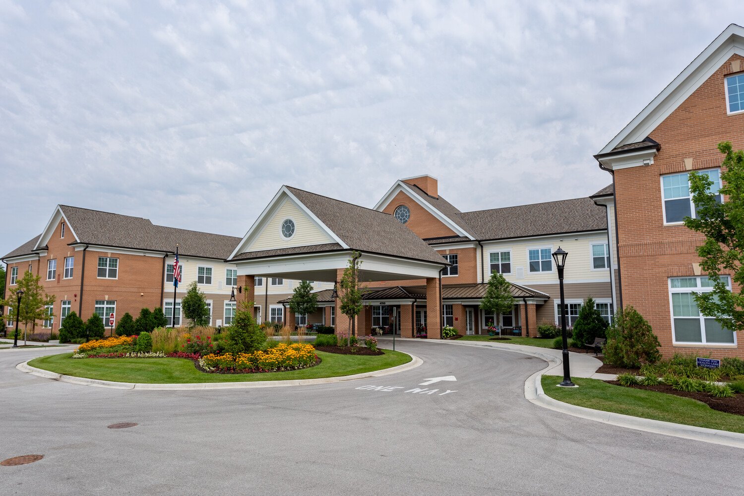 The Front of Arbor Terrace Glenview with a roundabout carpark area. a grass area with bushes and flowers. a brick and cream buidling behind.
