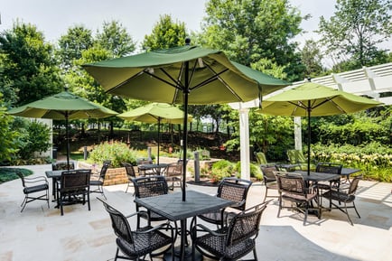 A beautifully landscaped outdoor patio with shaded dining tables, green umbrellas, and lush garden views at Arbor Terrace Herndon, offering the serene, resort-style independent living Herndon experience of a premier senior living Herndon Virginia community.
