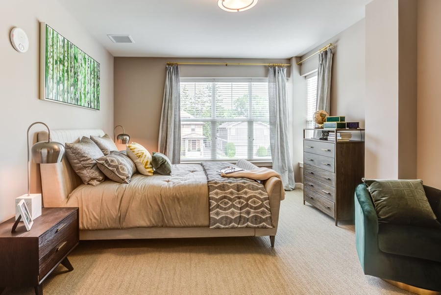 A model bedroom at Arbor Terrace Highland Park features a tufted bed with layered neutral and patterned bedding, a large window with soft curtains, a dark wood dresser, and a teal accent chair. It reflects the senior apartments near downtown Highland Park and senior living in Highland Park, IL.