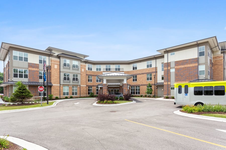 The brick and stone exterior of Arbor Terrace Highland Park features a covered entrance, landscaped grounds, a circular drive, and the community's accessible shuttle bus parked out front. This building is home to senior living and 55+ senior apartments on the North Shore.