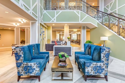 A two-story lobby at Arbor Terrace Highland Park features a grand staircase, blue floral armchairs around a wood coffee table, and an open reception and dining area beyond. It reflects the resort-style senior living in Lake County, IL, and senior living in Highland Park, IL.