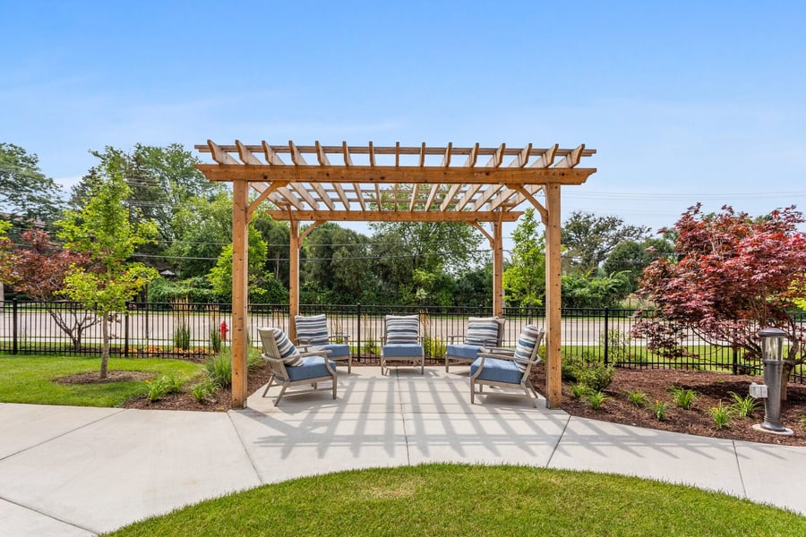 A wooden pergola shades cushioned outdoor seating at Arbor Terrace Highland Park, surrounded by landscaped gardens with shrubs and green trees under a clear blue sky. This peaceful courtyard reflects the nature-friendly senior living with walking paths in Highland Park, IL.