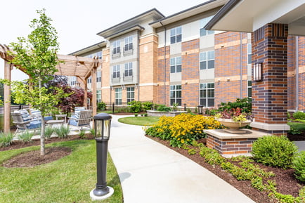 A landscaped courtyard at Arbor Terrace Highland Park features a wooden pergola, patio seating, and a paved path lined with yellow blooms, leading to the brick community building. It reflects the nature-friendly senior living with walking paths in Highland Park, IL.