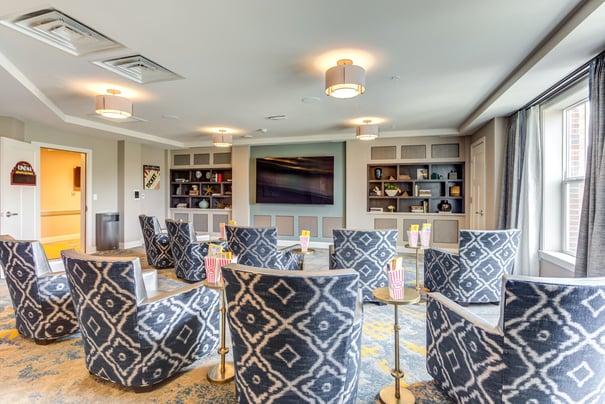 A cozy in-house cinema room at Arbor Terrace Highland Park features blue patterned armchairs, a large wall-mounted screen, built-in bookshelves, and popcorn boxes on gold side tables. This amenity reflects the resort-style senior living residents enjoy in Highland Park, IL.