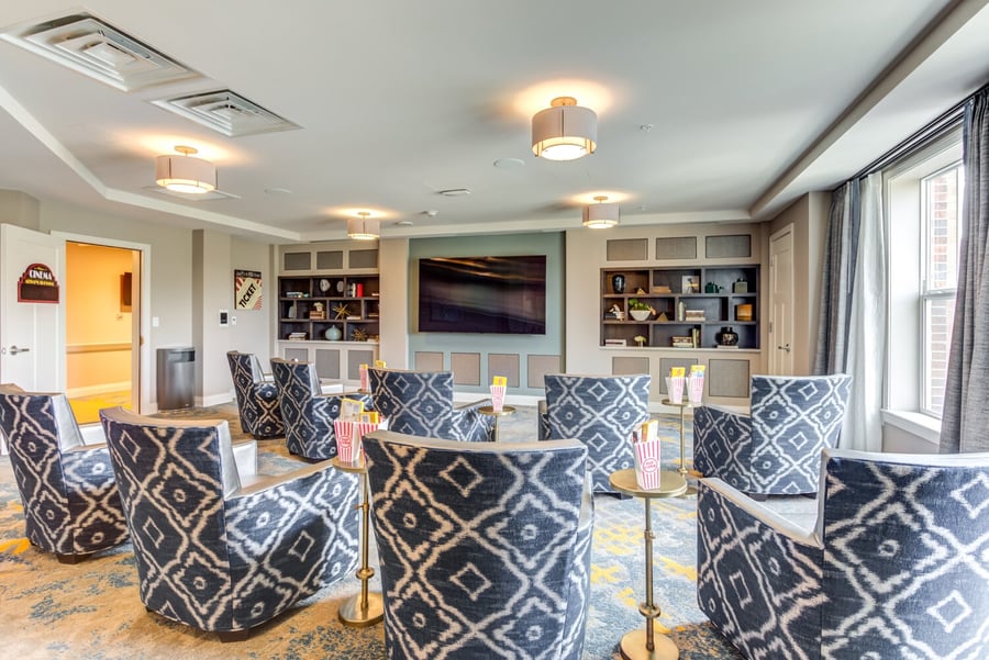 A cozy in-house cinema room at Arbor Terrace Highland Park features blue patterned armchairs, a large wall-mounted screen, built-in bookshelves, and popcorn boxes on gold side tables. This amenity reflects the resort-style senior living residents enjoy in Highland Park, IL.