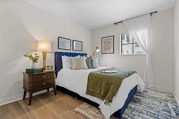 A bright bedroom with a navy upholstered bed, crisp white linens, an olive fringed throw, and a wood nightstand topped by a gold lamp and greenery. Senior apartments in Fort Worth, TX like this one at The Vantage at City View show how comfortable senior living in Fort Worth, TX can feel.