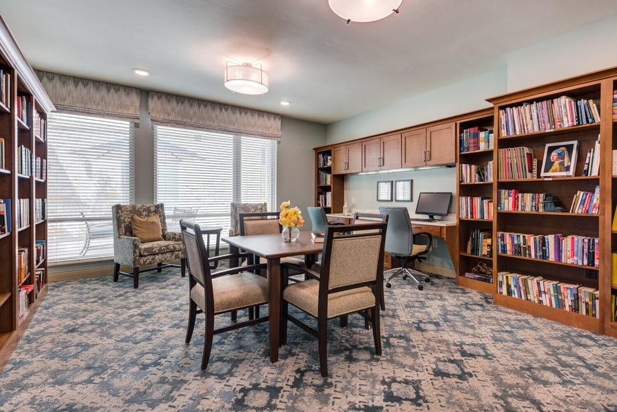 A bright, well-appointed library room with floor-to-ceiling bookshelves, a central table, and comfortable seating — thoughtful design inside a memory care community Lakeway TX.