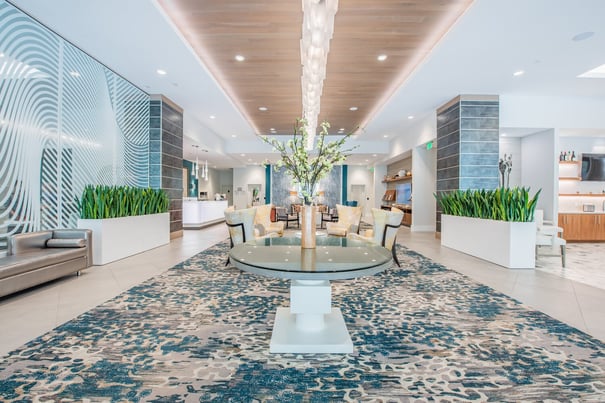 The welcoming grand lobby at Mirabelle in Miami, FL features a cascading chandelier, a floral centerpiece, lush greenery, and sophisticated seating, creating a warm first impression for senior living guests.