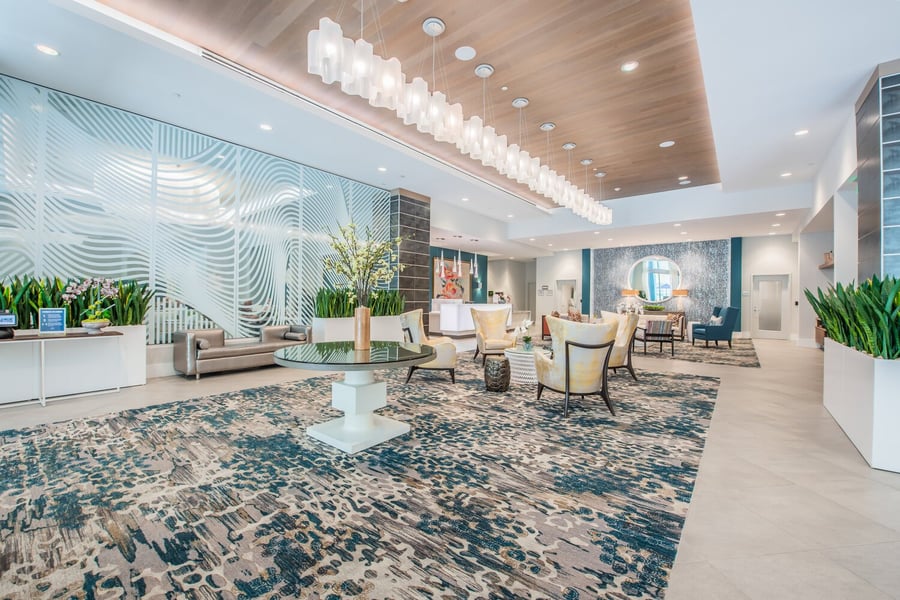The elegant lobby at Mirabelle features a dramatic chandelier, sculptural wall details, lush greenery, and stylish seating, setting a refined and welcoming tone for assisted living in Miami, FL.