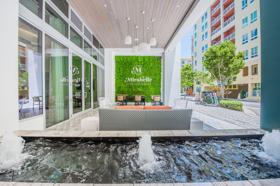 The inviting entrance of Mirabelle in South Miami, FL features a lush green living wall with the community's logo, a water feature, and stylish outdoor seating, welcoming residents and guests warmly.