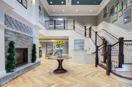 The elegant two-story lobby at Arbor Terrace Morris Plains features a stone fireplace, herringbone hardwood floors, a grand staircase, and fresh sunflowers, reflecting the upscale senior living experience in Morris Plains, NJ.