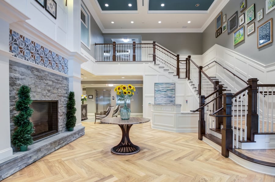 The stunning two-story foyer at Arbor Terrace Morris Plains showcases a stone fireplace, herringbone hardwood floors, and a sweeping staircase, embodying the warm, welcoming spirit of senior care in Morris Plains, NJ.