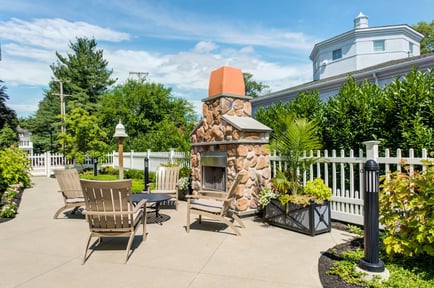 A charming outdoor patio at Arbor Terrace Morris Plains features a stone fireplace, Adirondack chairs, and lush garden surroundings, offering a peaceful nature-focused senior living experience in Morris Plains, NJ.