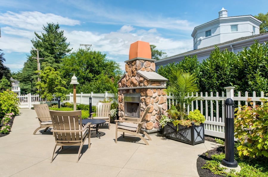 A charming outdoor patio at Arbor Terrace Morris Plains features a stone fireplace, Adirondack chairs, and lush garden surroundings, offering a peaceful nature-focused senior living experience in Morris Plains, NJ.