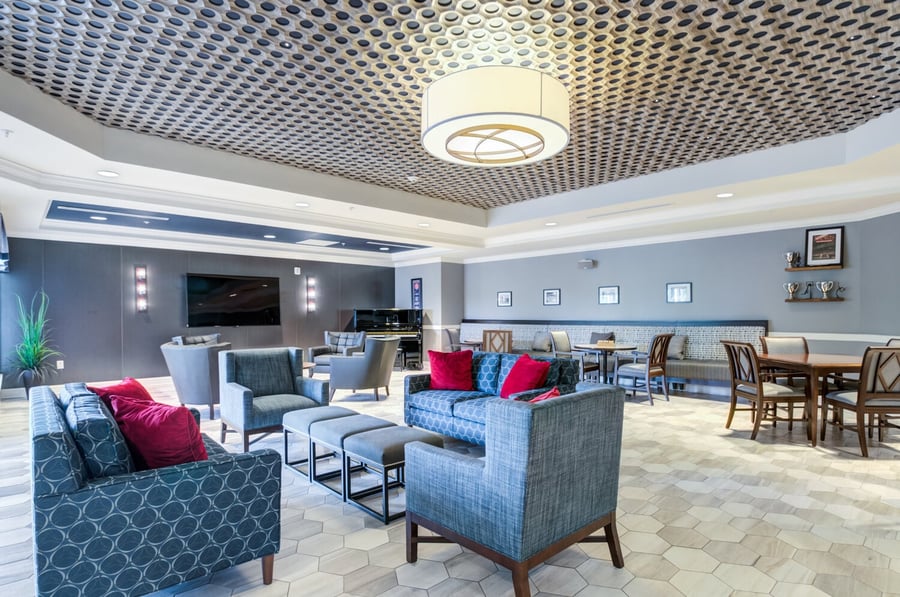 A spacious, elegantly designed common lounge at Arbor Terrace Morris Plains features stylish seating, bold accent pillows, a large TV, and a patterned ceiling, showcasing the upscale assisted living community in Morris Plains, NJ.