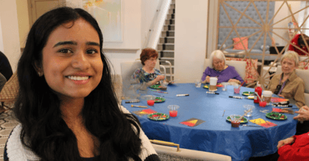 A smiling person in the foreground joins residents gathered around a colorful activity table for a creative arts session, showcasing the vibrant, engaging lifestyle at our senior living East Cobb community.