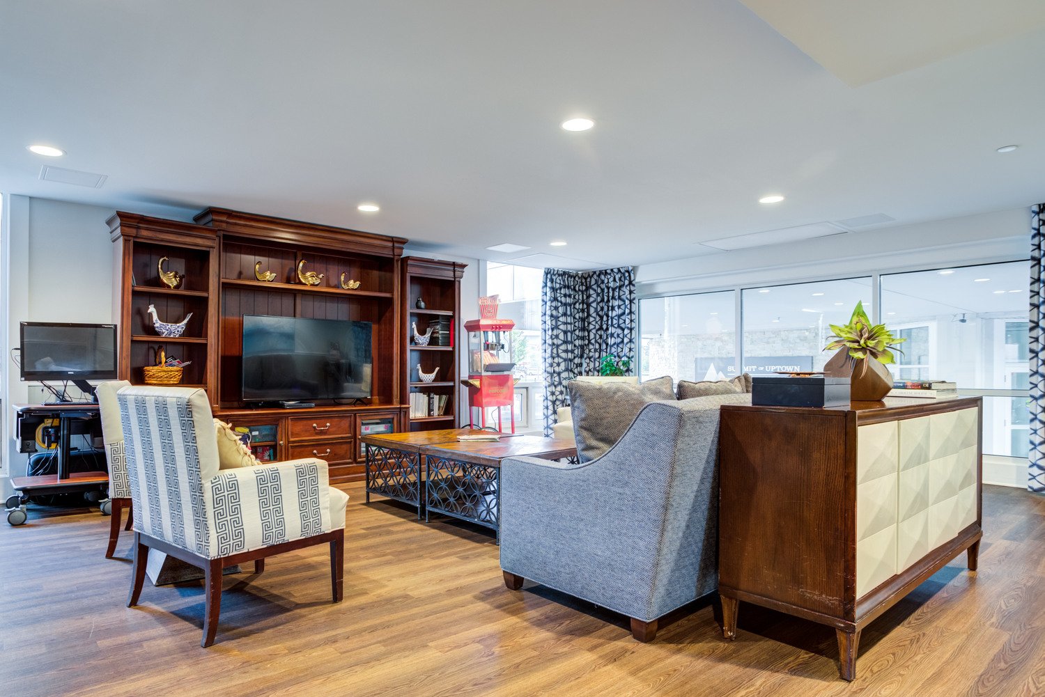 Summit of Uptown common space with a grey couch backing up to a table. Black and white striped chairs next to the couch and a coffee table in the center. tv in the front of the room on wooden shelving. Big window with curtains to the side of the room.