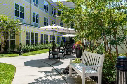A serene outdoor courtyard with shaded patio seating, lush trees, and colorful flowers surrounding the grounds of our senior living community in Gambrills, MD.